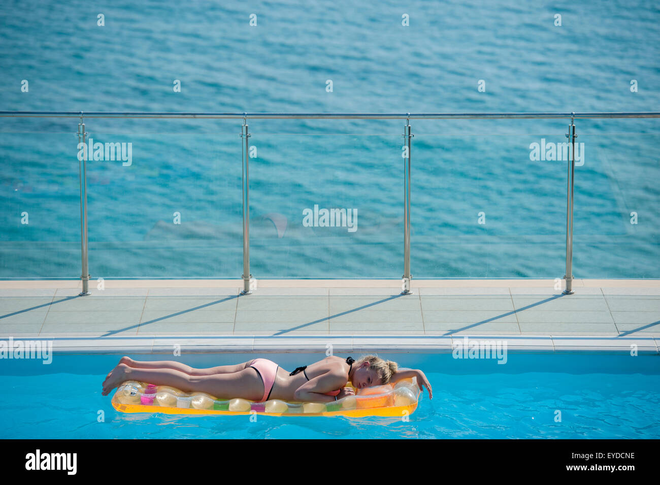Una hermosa rubia descansa sobre una colchoneta inflable en una piscina con el azul del mar directamente detrás de ella Foto de stock