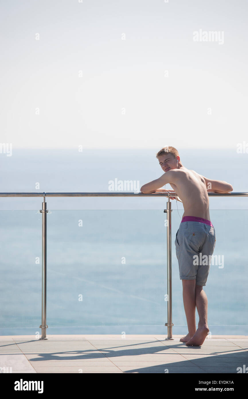 Un adolescente está en la piscina y se inclina contra una pared de vidrio mirando al mar debajo Foto de stock