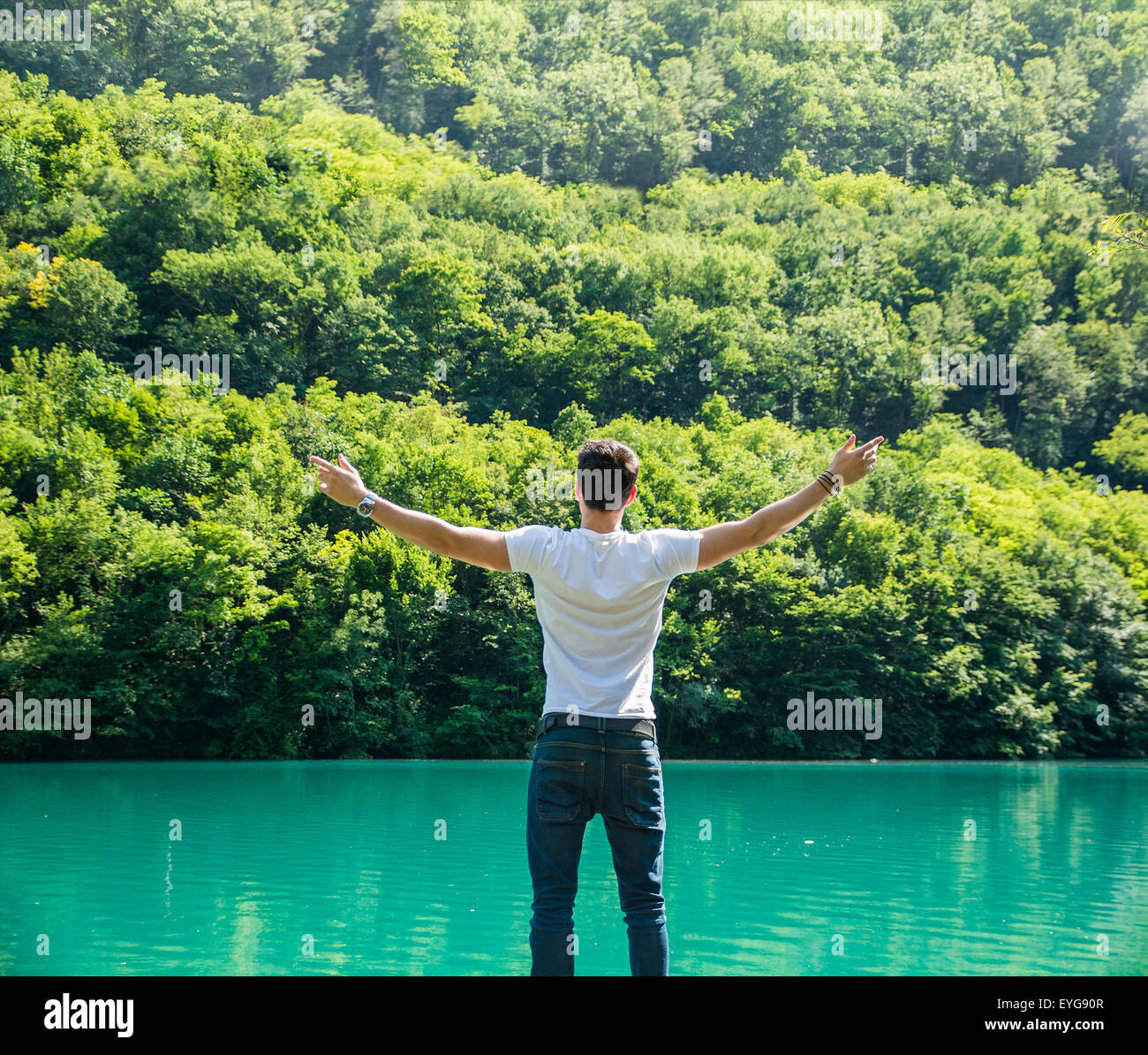 Joven en camiseta, con los brazos abiertos al aire libre disfrutando de libertad frente al lago, visto desde la parte posterior. Foto de stock