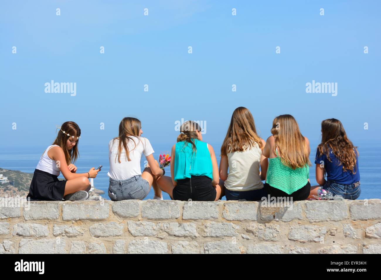 Grupo de chicas adolescentes charlando, sentándose juntas en una pared y mirando al mar Foto de stock