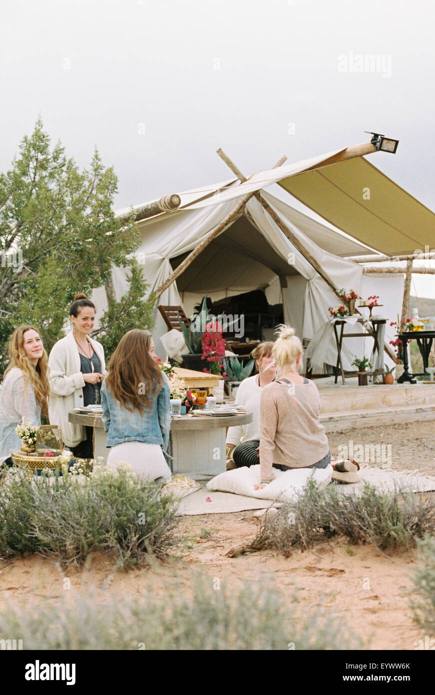 Grupo de amigas, sentados en el suelo alrededor de una mesa en un desierto, una carpa en el fondo. Foto de stock