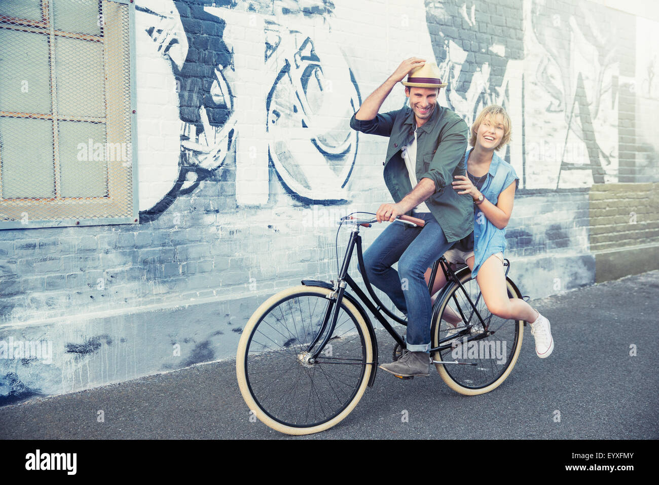 Retrato pareja juguetona montando bicicleta por urban graffiti wall Foto de stock