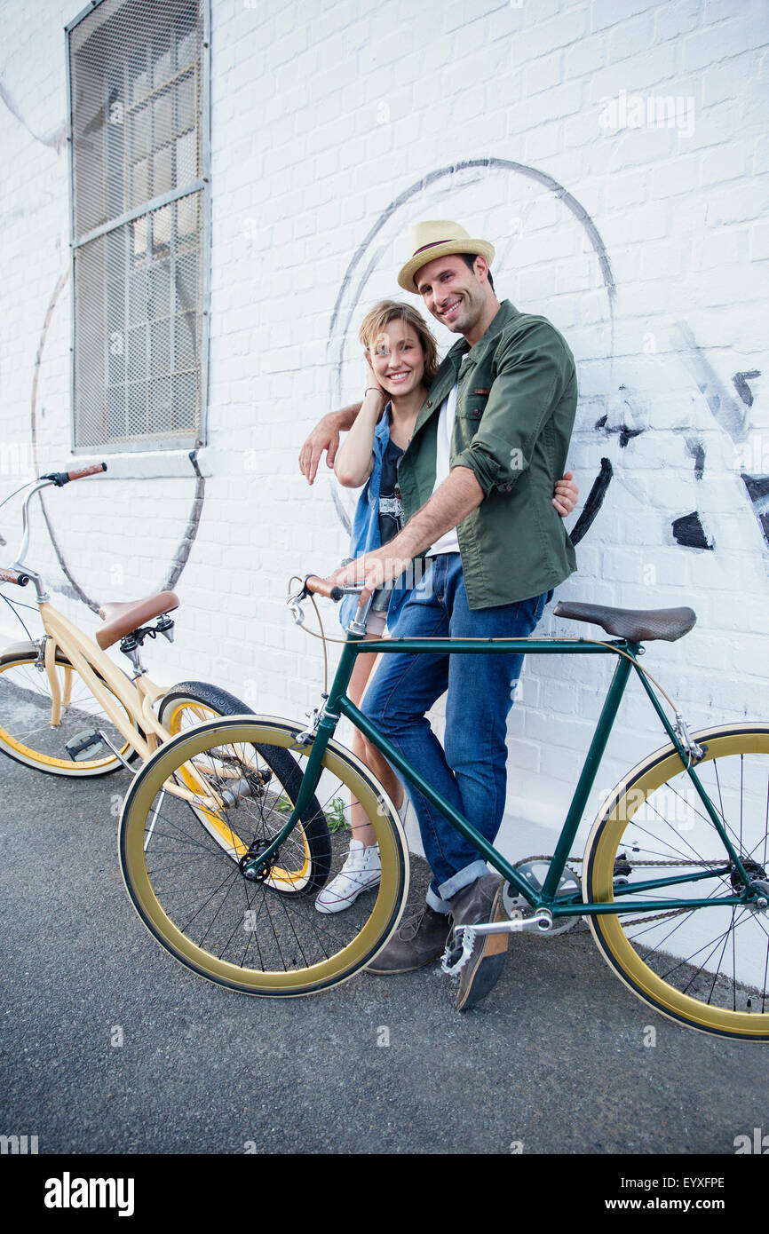 Retrato sonriente pareja con bicicletas abrazando a pared urbana Foto de stock