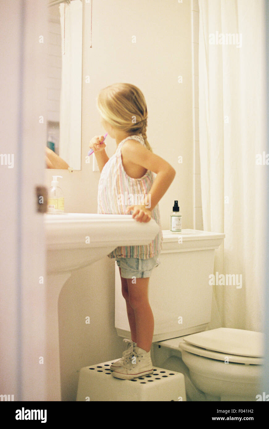 Niña de pie sobre un taburete en el cuarto de baño, cepillando sus dientes. Foto de stock