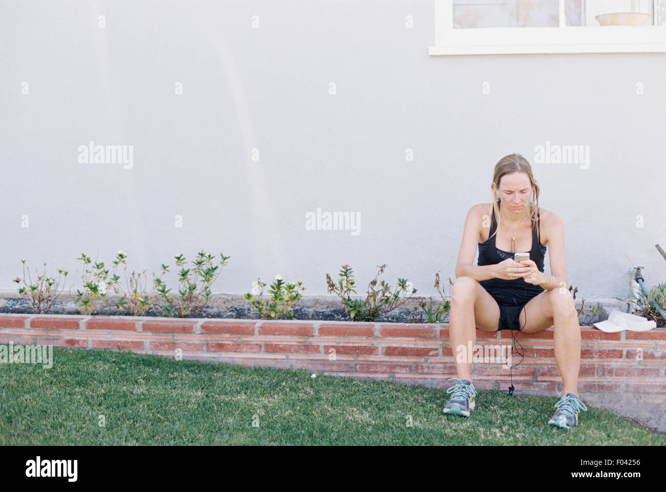 Mujer rubia en ropa deportiva sentados en el muro de un jardín, descansando después de un jog. Foto de stock