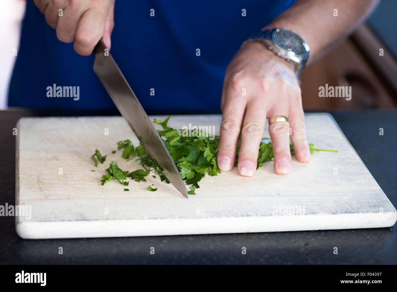 Un hombre cortando perejil fresco en rama para una comida en una tabla de cortar con un cuchillo de cocina grande Foto de stock