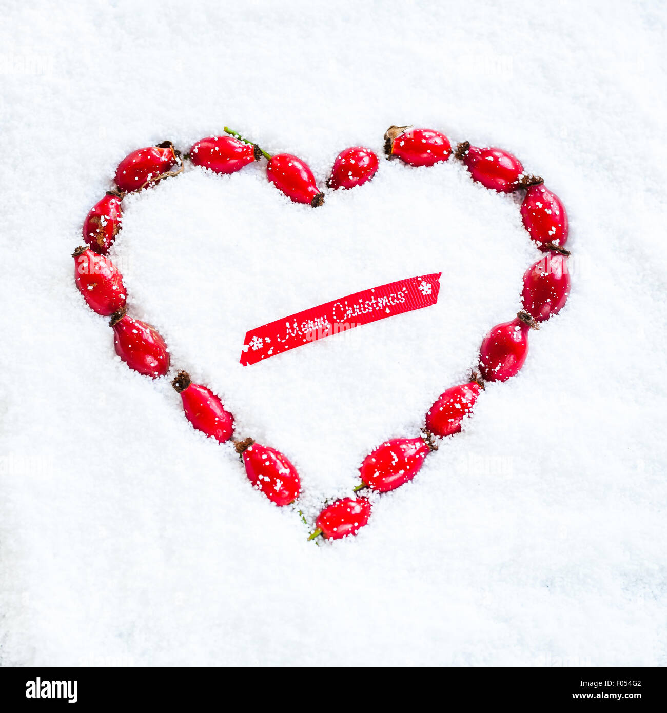 Una corona con forma de corazón hecho con escaramujos establecidos en la nieve, y el trozo de cinta roja diciendo ¡Feliz Navidad! Foto de stock