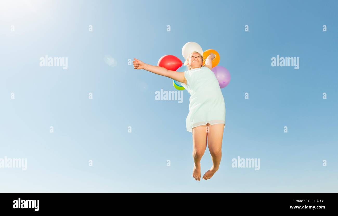 Chica sujetando ramo de globos saltando el aire contra el cielo azul Foto de stock