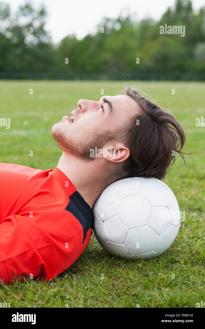 Vista lateral del joven cabeza descansando sobre una pelota de fútbol en el campo Foto de stock