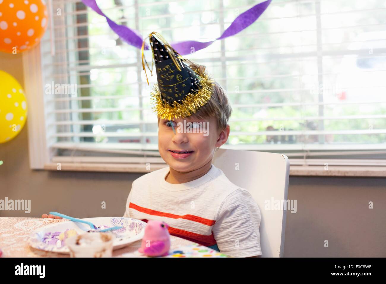 Muchacho de gorro de fiesta sentado en una mesa comiendo pastel de cumpleaños tirando cara divertida Foto de stock