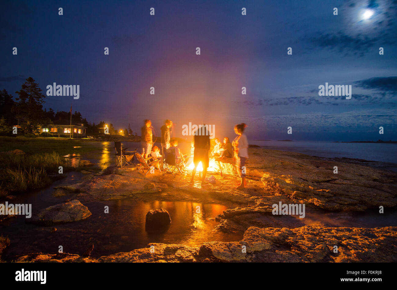 La familia y los amigos por una cálida fogata bajo una luna iluminada de noche como marea alta escurre lentamente hacia el interior. Foto de stock