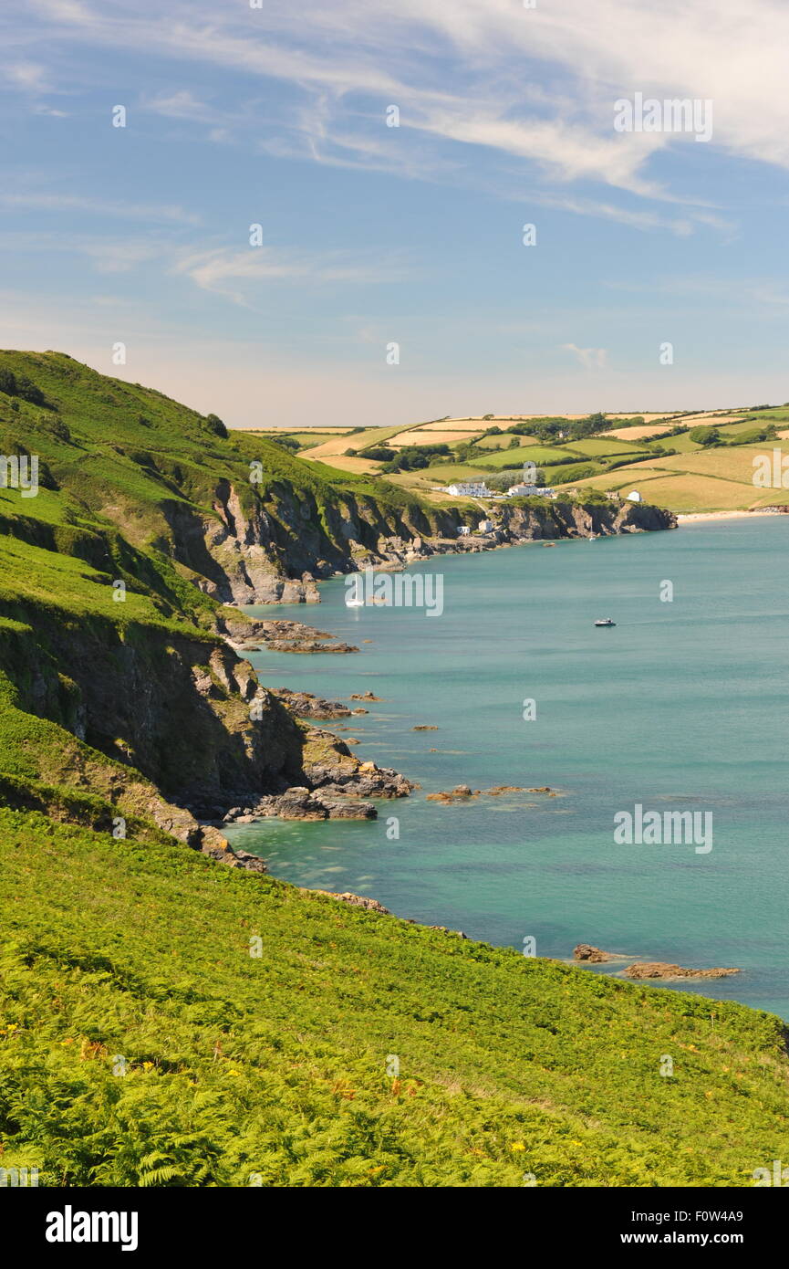 Soleada costa Inglesa, mar calma con alta tenues nubes y campos verdes. Velero y lejano pueblo costero. Foto de stock