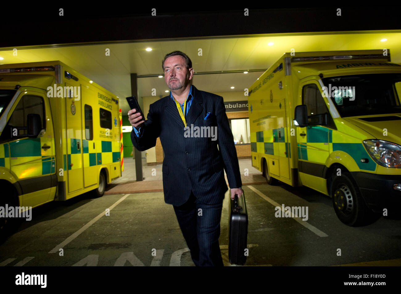 Un hombre fuera de horario, doctor en llamada, manteniendo la bolsa Médica y teléfono móvil, pasando por una línea de ambulancias en un hospital UK GP Foto de stock