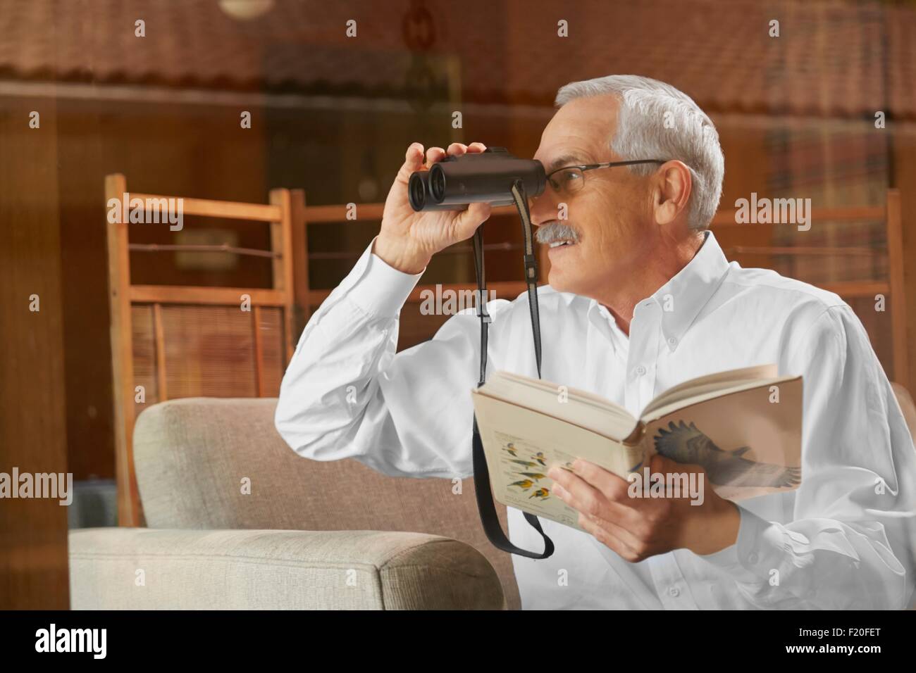 Hombre senior en casa, a través de la ventana de observación de aves, utilizando binoculares Foto de stock