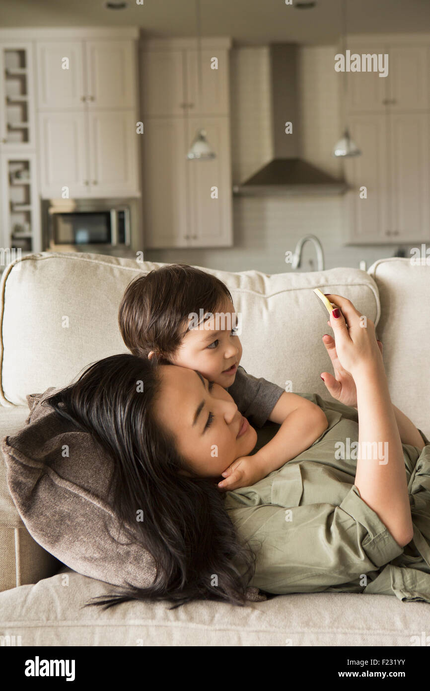 Mujer recostada sobre un sofá caricias con su joven hijo y mirando un teléfono celular. Foto de stock