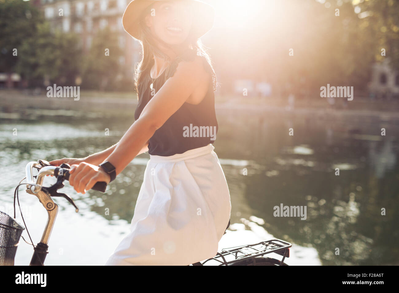 Retrato de mujer bastante joven montando en su bicicleta mirando a otro lado, sonriente, con la llamarada solar. Mujer ciclismo en un día de verano. Foto de stock