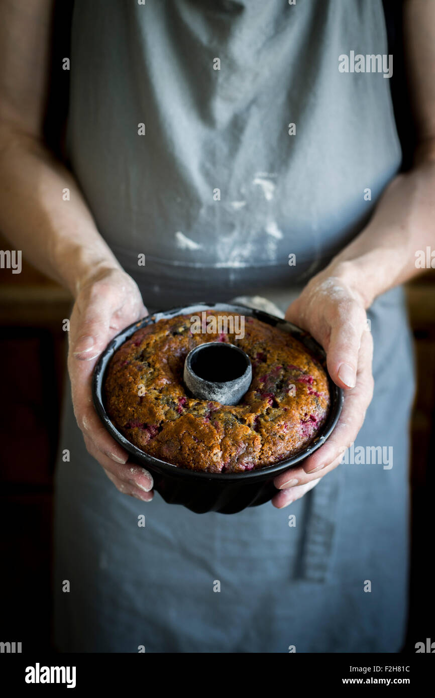 Persona con delantal de cocina y manos sosteniendo enharinada bundt cake en la bandeja de hornear Foto de stock