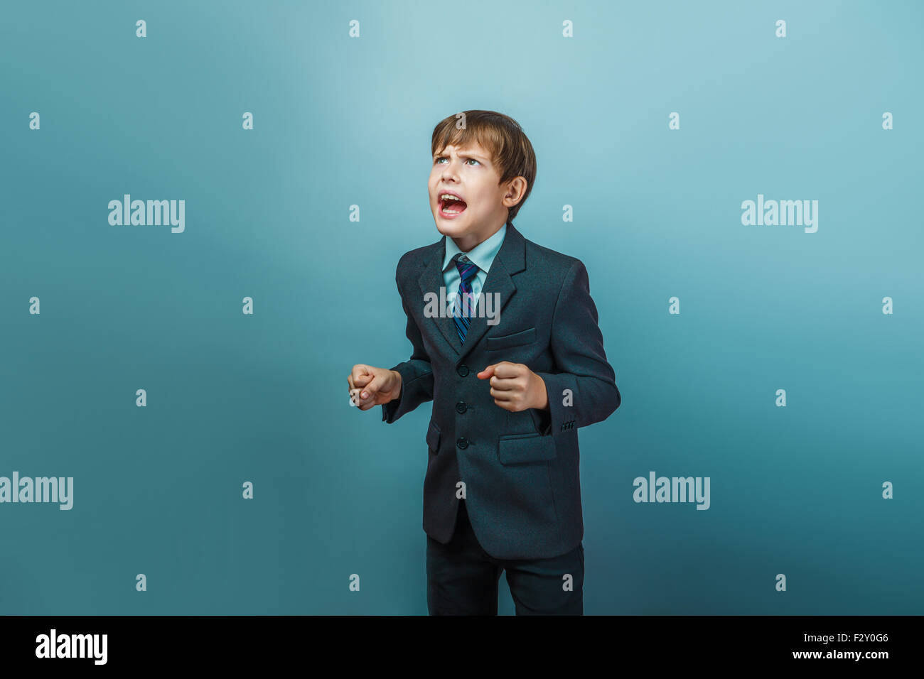 Un muchacho de doce en un traje de apariencia europea gritando enojado sobre un fondo gris Foto de stock