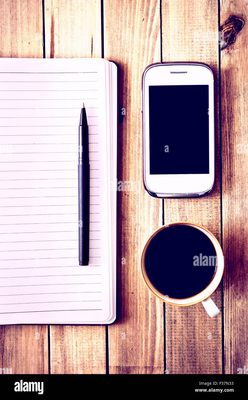Teléfono celular blanco, un bolígrafo, una taza de café y el portátil en la mesa de madera. Espacio de trabajo. Vintage Instagram fotografía. Foto de stock