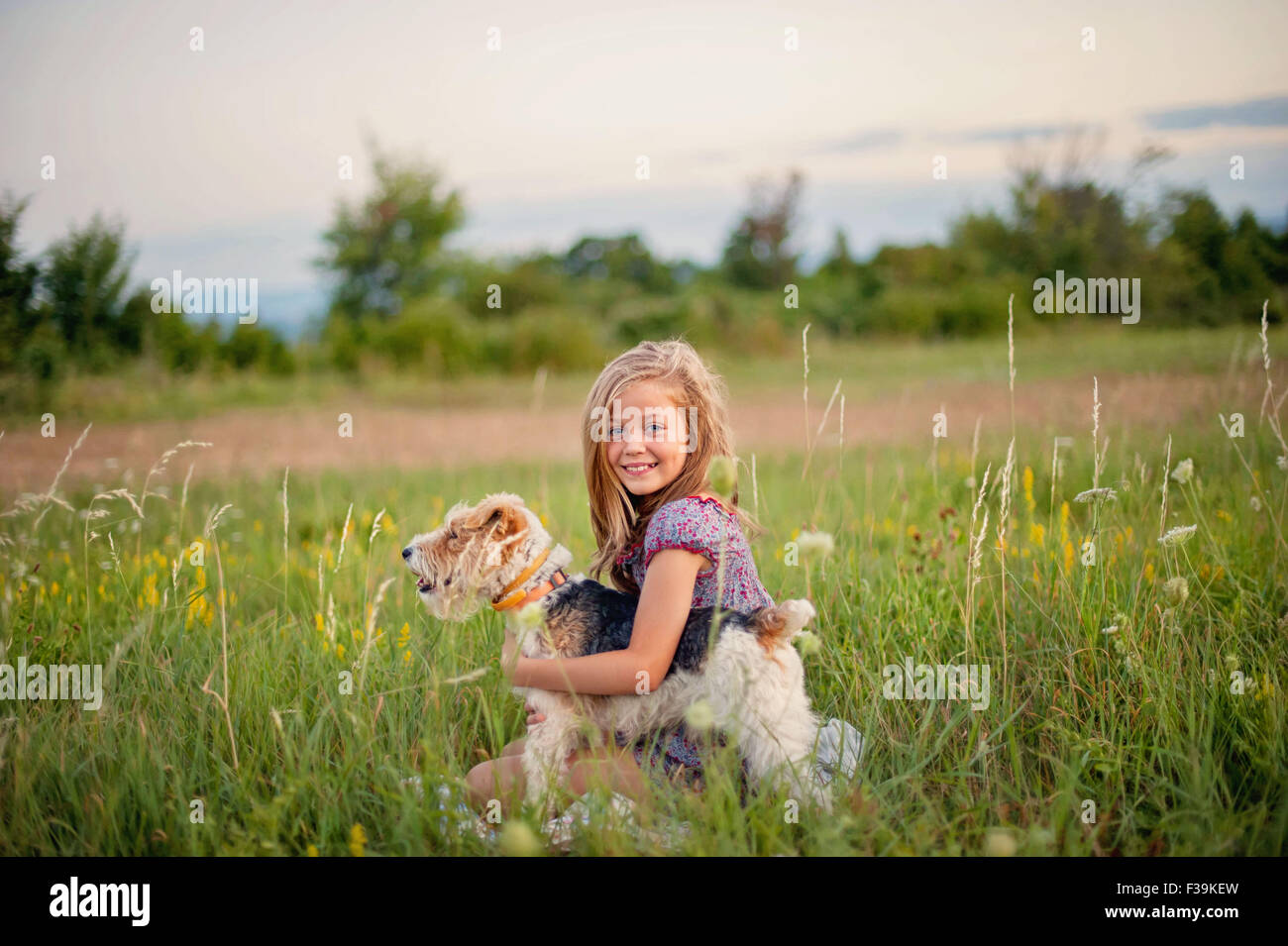 Retrato de una sonriente joven sentada en el campo abrazando a su perro Fox Terrier Foto de stock
