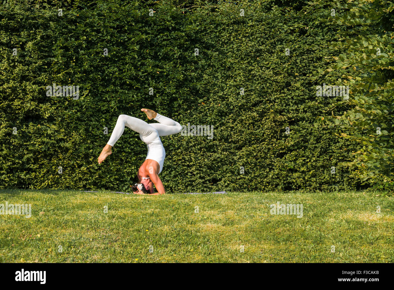 Mujer joven, vistiendo un traje de cuerpo blanco, es practicar el Hatha-yoga piscina entre árboles, mostrando la pose: sirshasana, soporte de cabezal Foto de stock