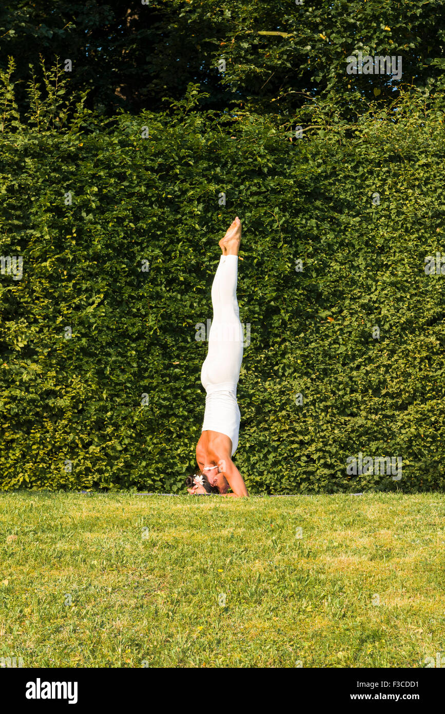 Mujer joven, vistiendo un traje de cuerpo blanco, es practicar el Hatha-yoga piscina entre árboles, mostrando la pose: sirshasana, soporte de cabezal Foto de stock