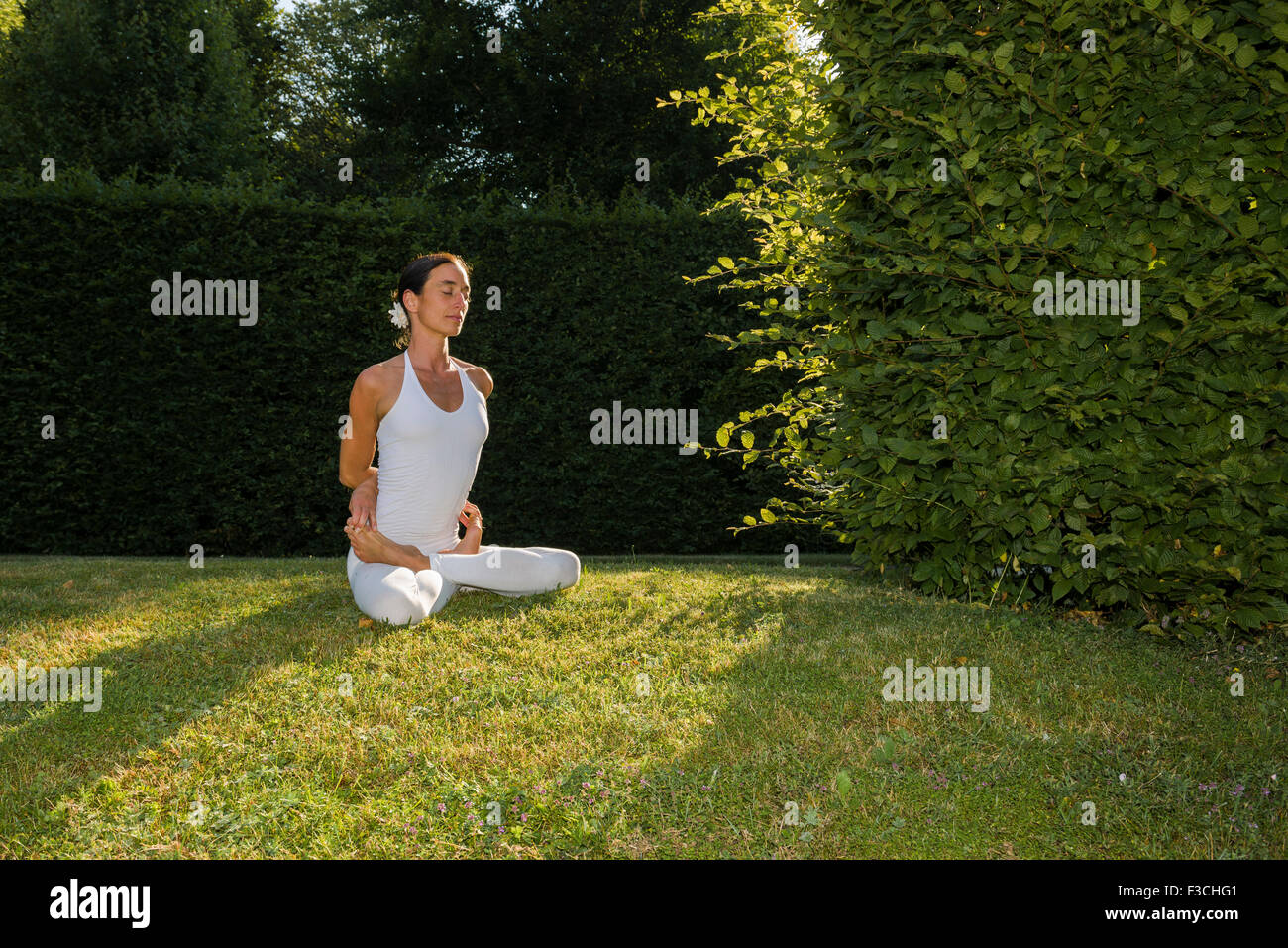 Mujer joven, vistiendo un traje de cuerpo blanco, es practicar el Hatha-yoga piscina entre árboles, mostrando la pose: padmasana, lotus plantean Foto de stock