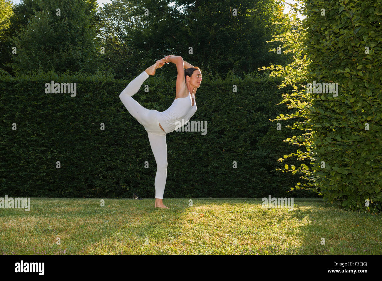 Mujer joven, vistiendo un traje de cuerpo blanco, es practicar el Hatha-yoga piscina entre árboles, mostrando la pose: natarajasana, señor shi Foto de stock