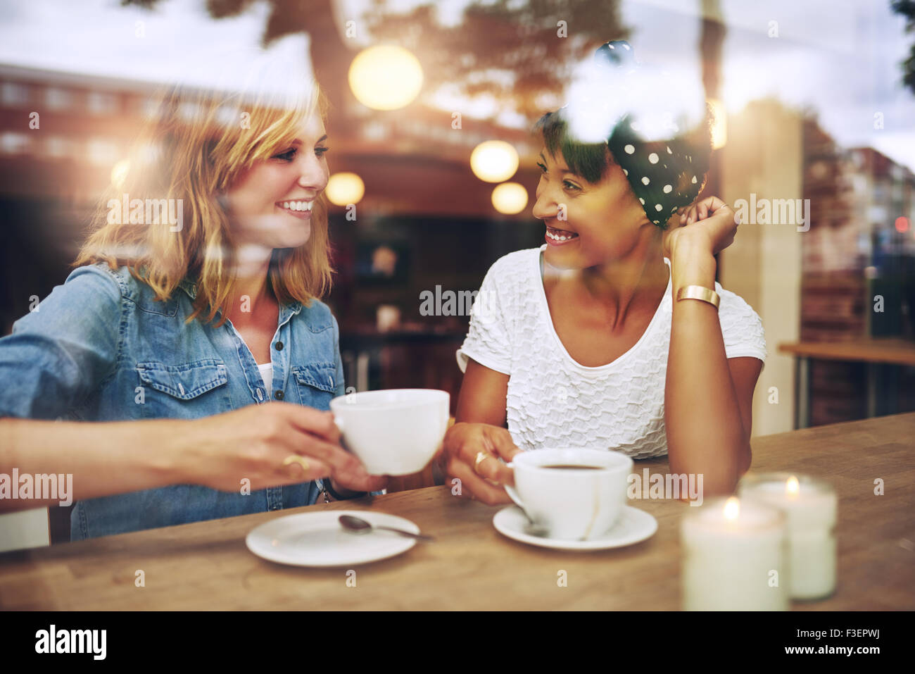 Dos amigos disfrutando multiétnico café juntos en una cafetería vistos a través del cristal con reflejos mientras se sientan en una mesa Foto de stock