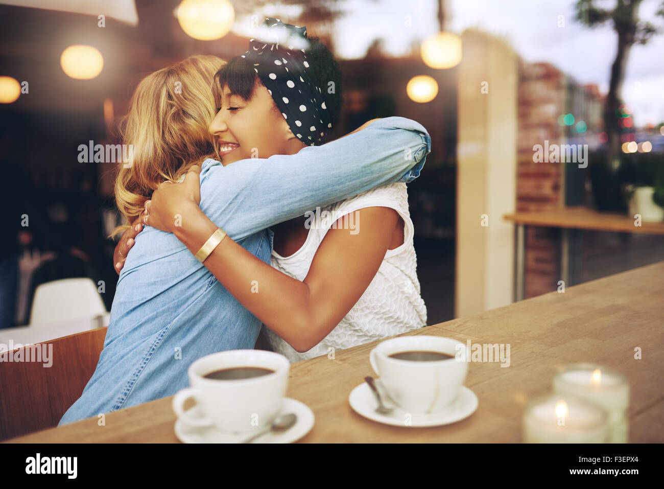Dos amigas pluriétnico afectuoso abrazo mientras se sientan en un café disfrutando de una taza de café juntos Foto de stock