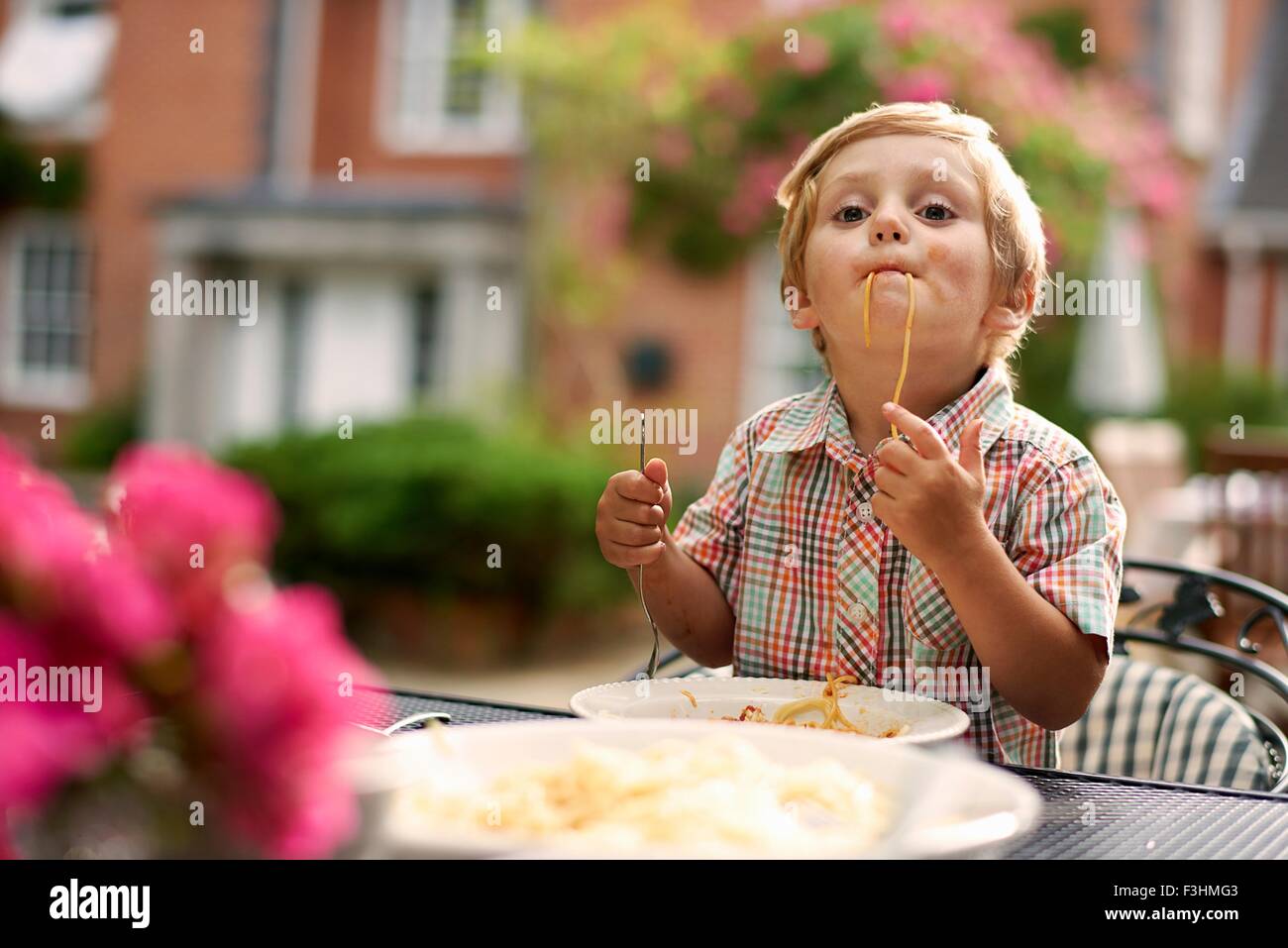 Muchacho sentado en la mesa de jardín chupando spaghetti mirando a la cámara Foto de stock