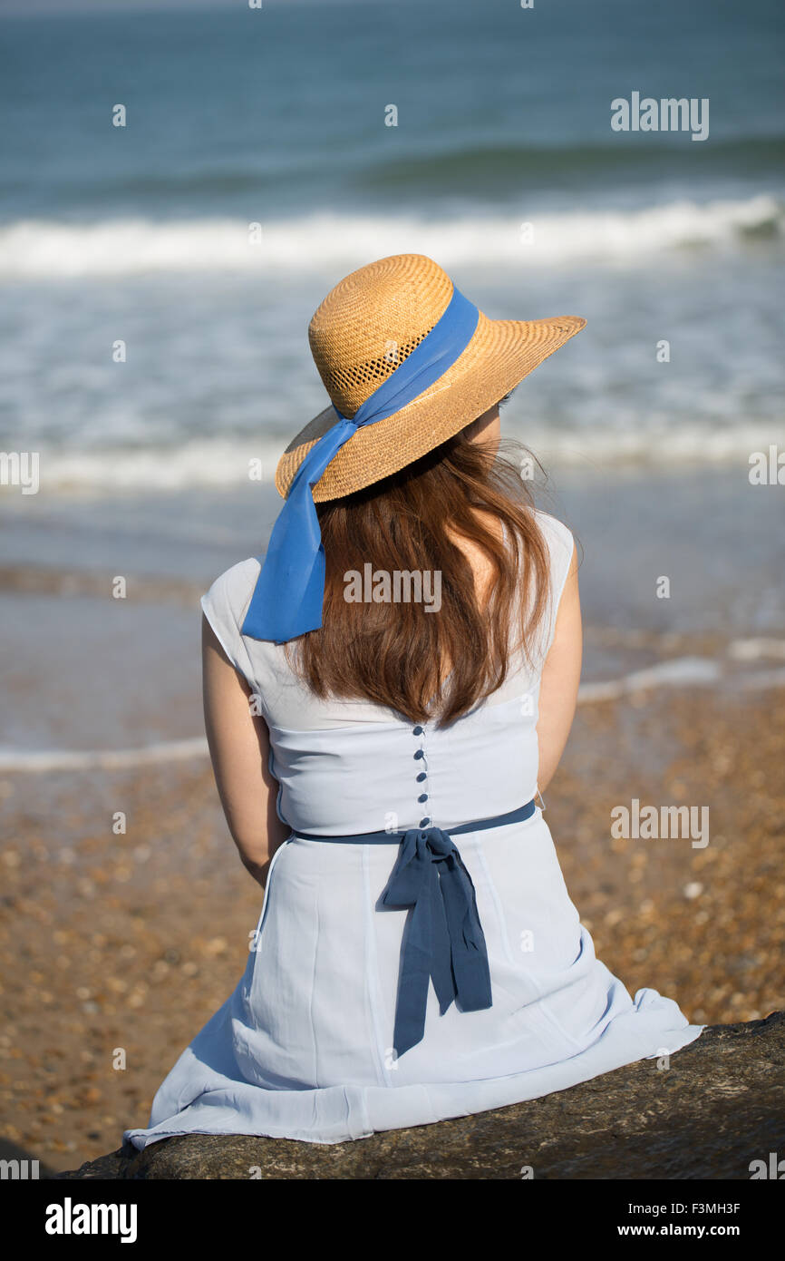 Joven y bella muchacha sentada histórico en la playa en un vestido azul y llevaba un sombrero de paja Foto de stock
