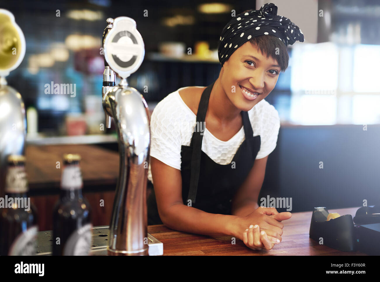 Atractiva joven dueño de la pequeña empresa americana africana vestida con un pañuelo y delantal recostada sobre el mostrador de su pub smili Foto de stock