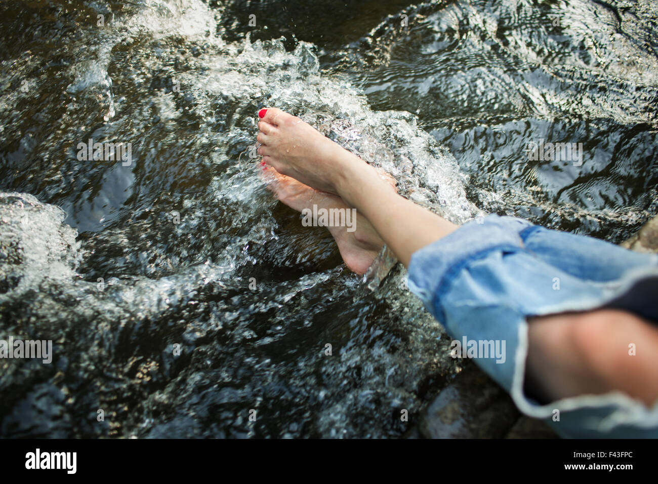 Una mujer en jeans de moda con un rip, con sus pies en las frías aguas de un río que fluye. Foto de stock