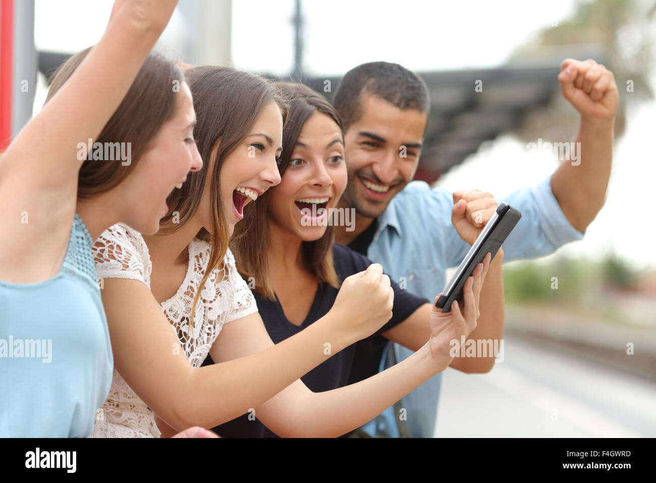 Cuatro eufóricos pluriétnico caucásicas y amigos árabes viendo la televisión en una tableta en una estación de tren Foto de stock