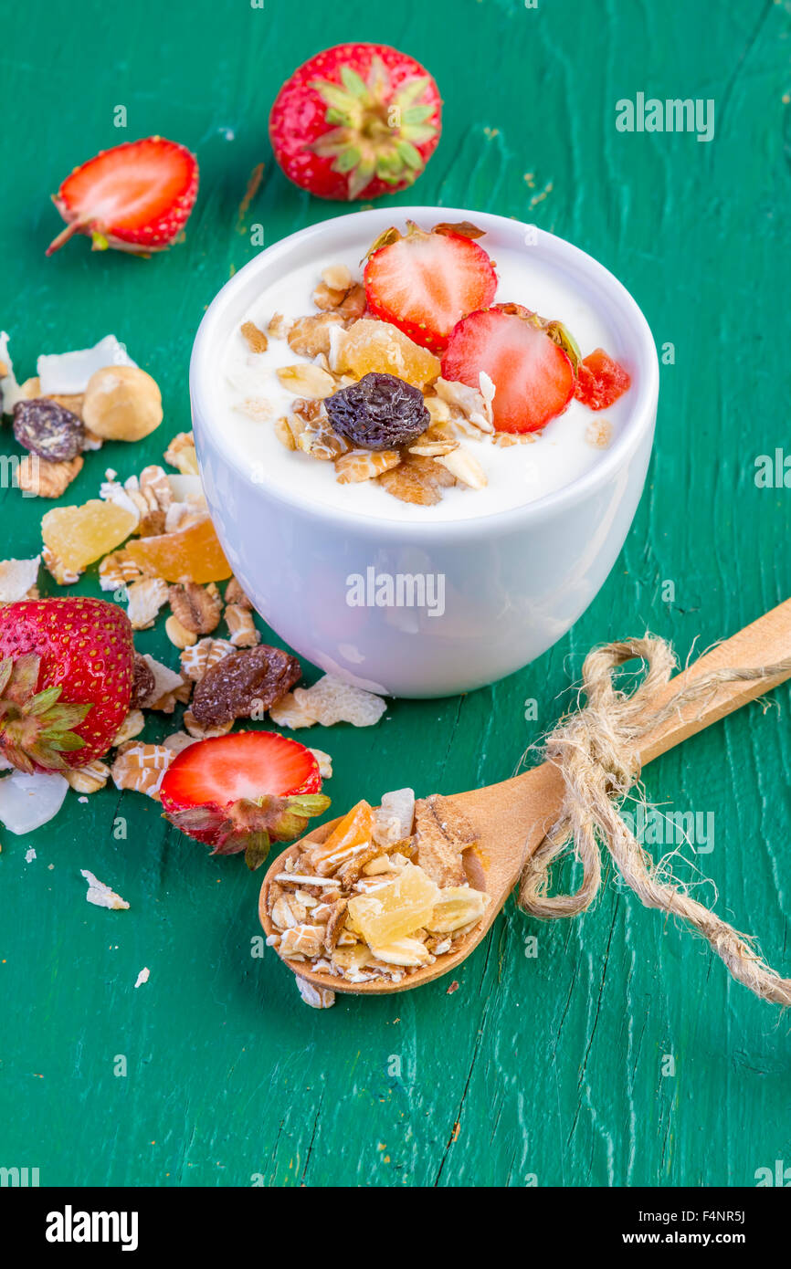 Yogur con cereales, muesli, fresas frescas, plátano y pasas en recipiente sobre fondo de madera Foto de stock