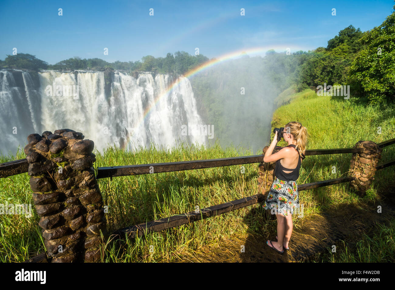 LIVINGSTONE, ZAMBIA - Mujer tomando fotografías de Victoria Falls cascadas con arco iris Foto de stock