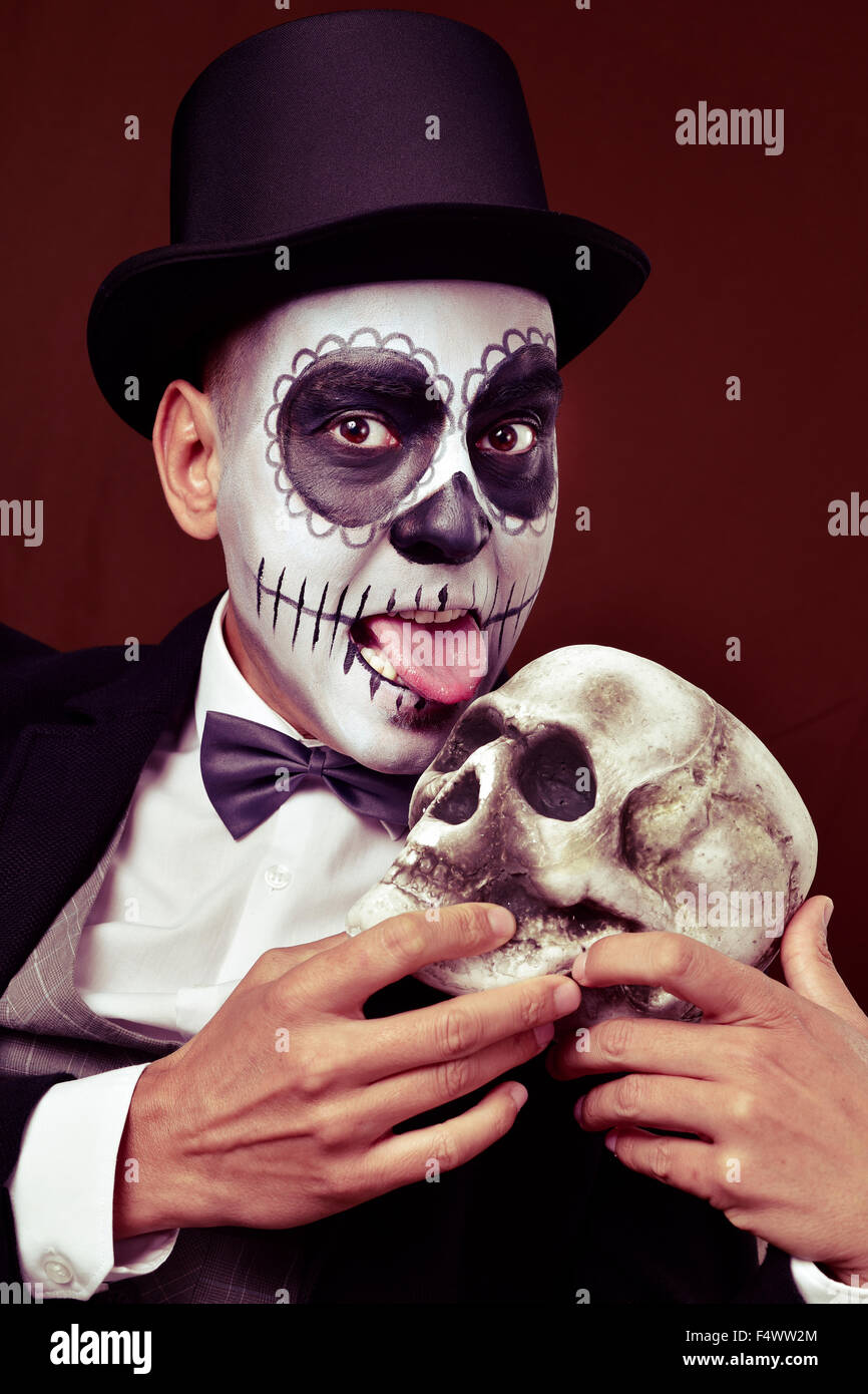 Un hombre joven con calaveras mexicanas maquillaje, vestido con sombrero de  copa y pajarita, lame un cráneo de miedo Fotografía de stock - Alamy