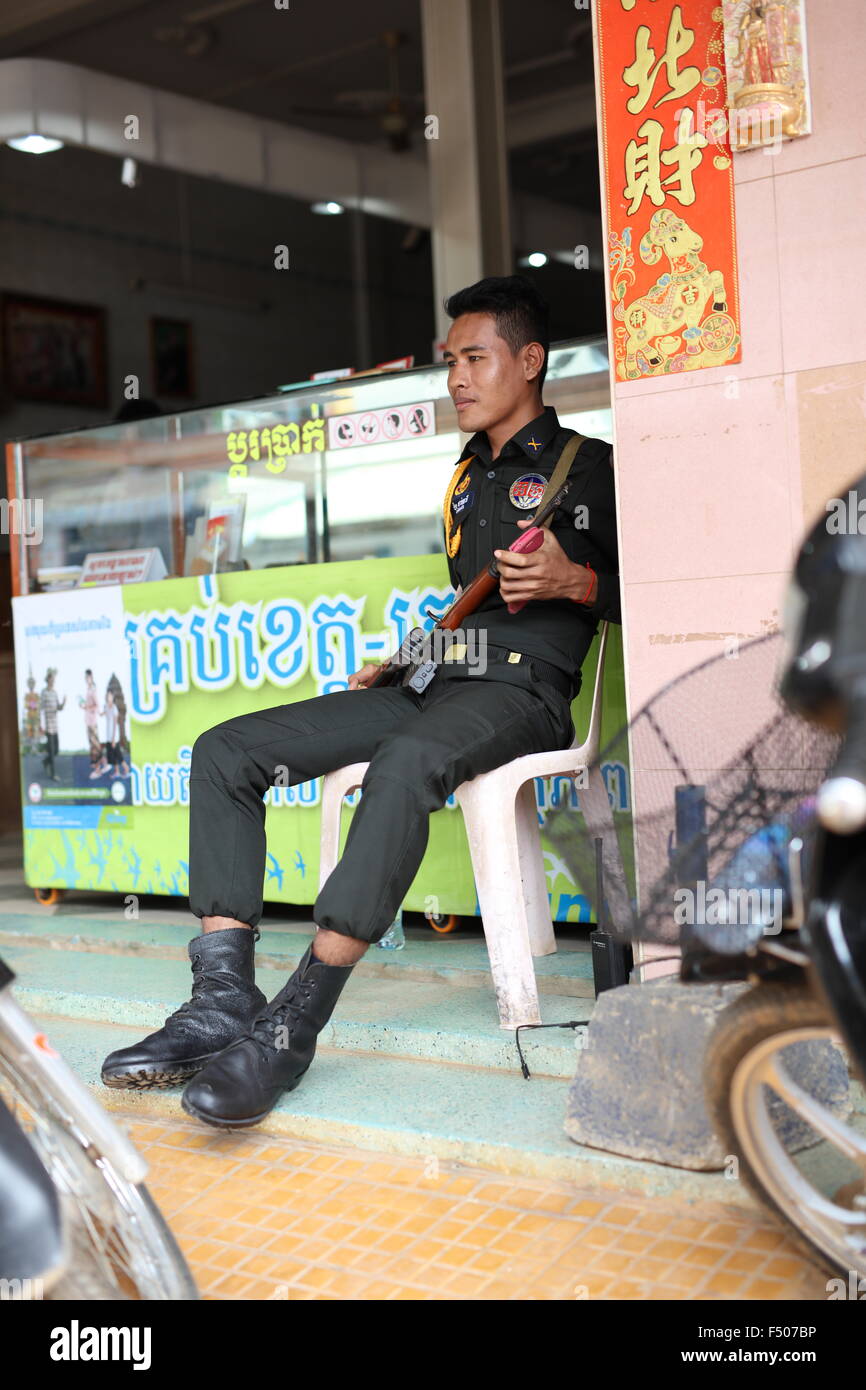 La policía hombre con fusil automático y teléfono móvil custodiando el cambio de dinero en Sisophon, Camboya Foto de stock