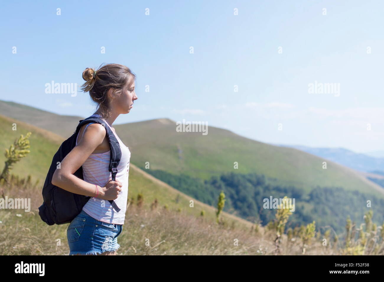 Joven, atractiva chica con una mochila en la espalda, de pie en la montaña. Praderas verdes y montañas majestuosas en el backgr Foto de stock