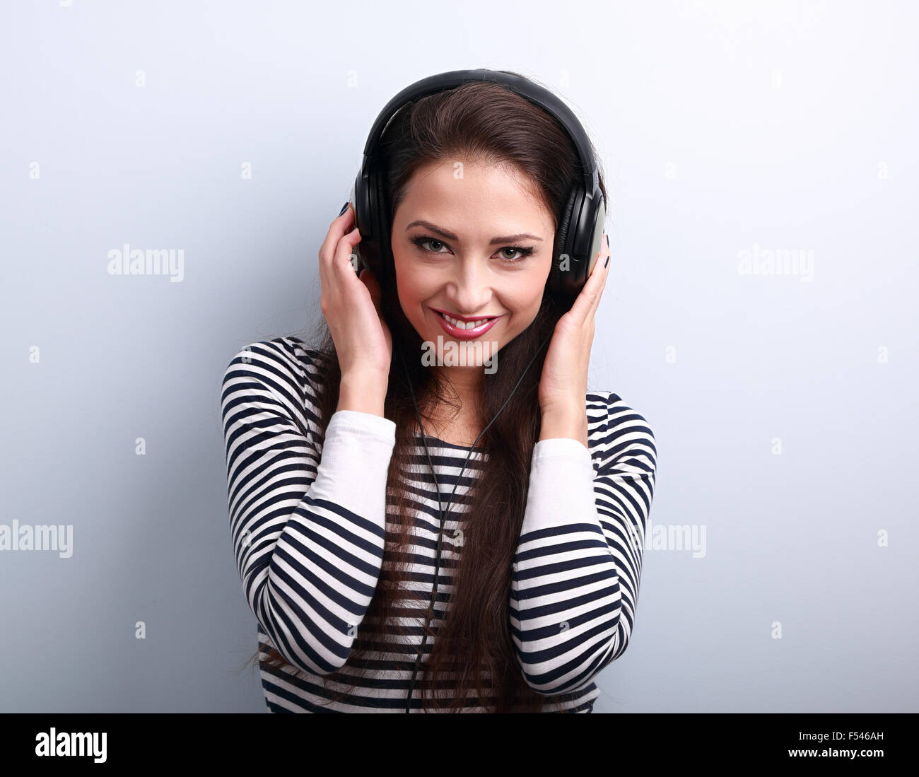 Feliz natural emoción mujer escuchando la música sosteniendo las manos auriculares y mirando sobre fondo azul. Foto de stock