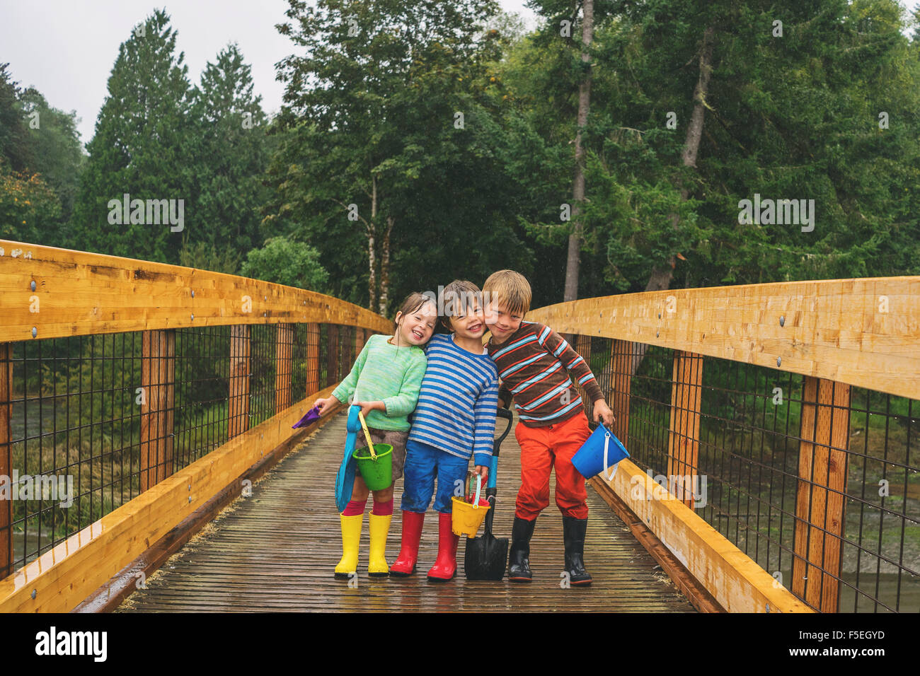 Dos chicos y una chica de pie sobre un puente de madera en la lluvia Foto de stock