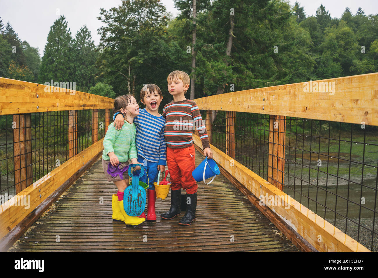 Dos chicos y una chica de pie sobre un puente de madera en la lluvia Foto de stock
