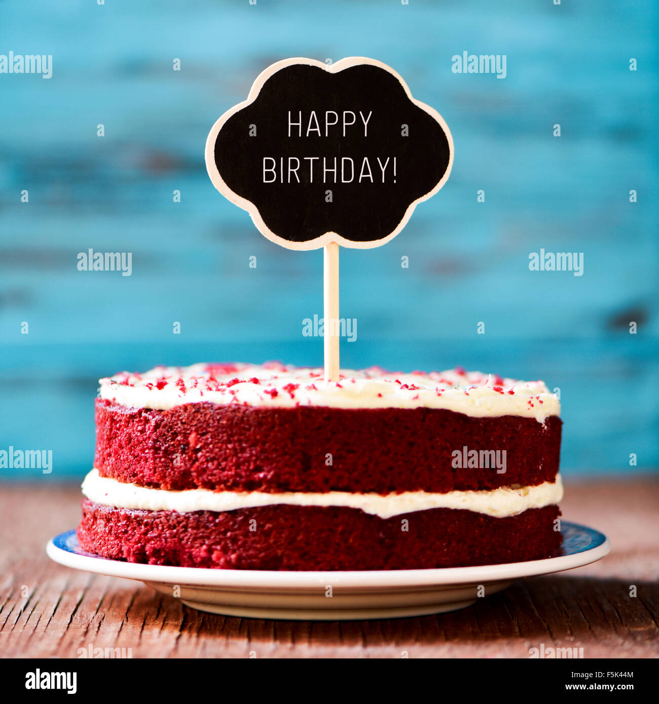 Un pastel de terciopelo rojo con una pizarra en la forma de un pensamiento de burbuja con el texto ¡Feliz cumpleaños, sobre una tabla de madera rústica Foto de stock