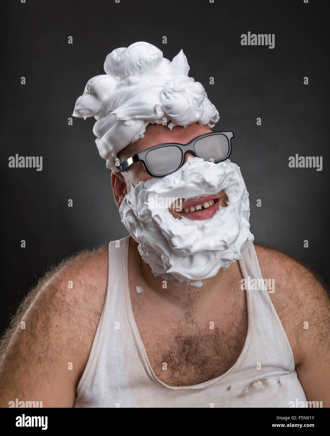 Extraño hombre sonriente en gafas con espuma de afeitar en su rostro y en su cabeza sobre fondo gris Foto de stock