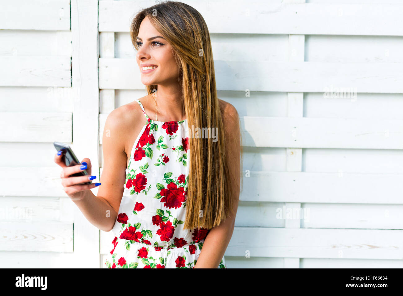 Hermosa joven utilizando el teléfono mientras se apoya contra una pared blanca durante un brillante día de verano Foto de stock