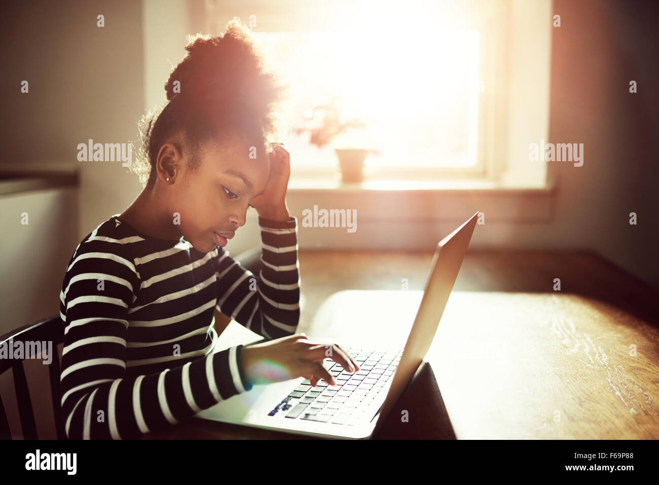 Joven negro niña trabajando en sus deberes en casa escribiendo información en un ordenador portátil, vista lateral contra un sol brillante resplandor Foto de stock