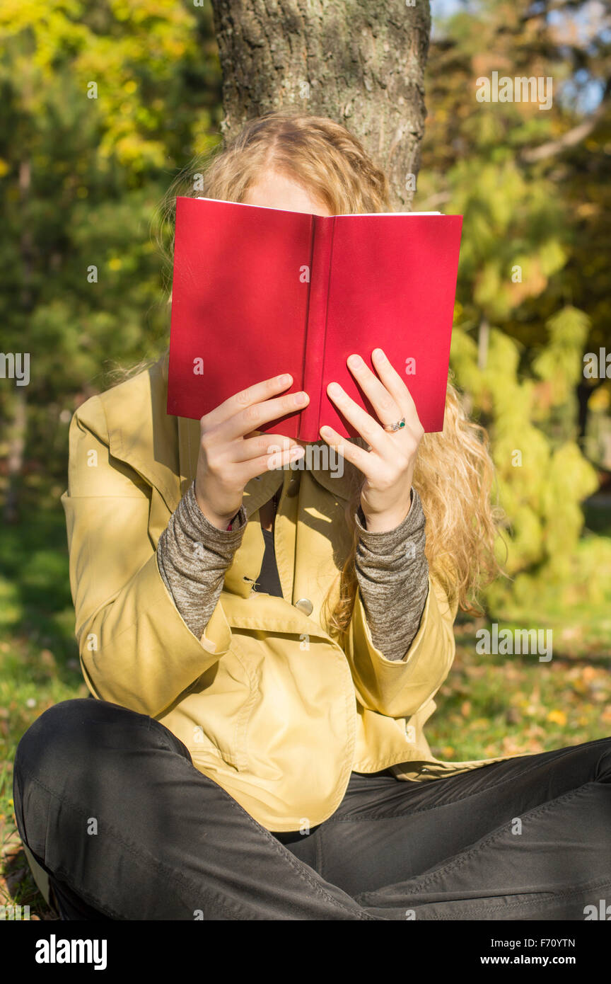 Chica rubia leyendo un libro rojo en un parque en un soleado día de otoño Foto de stock