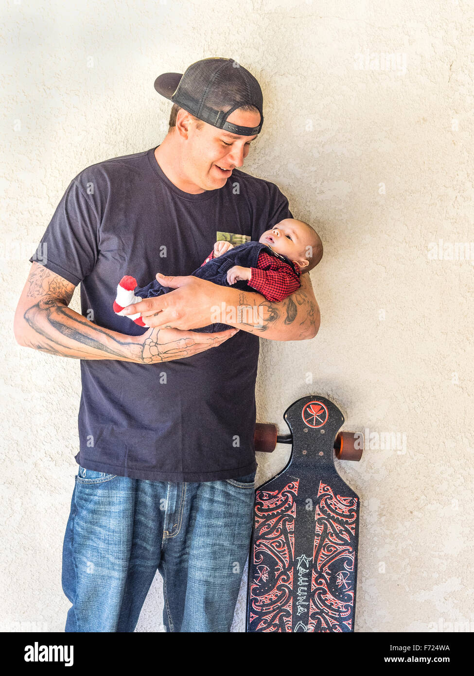 Skateboarding padre milenario con tatuajes 2 1/2 mes sosteniendo a su hijo y de pie junto al papá de la patineta. Foto de stock