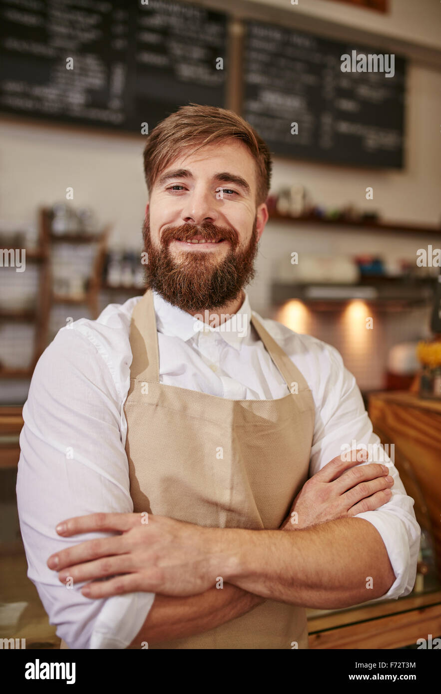 Retrato de joven feliz barista en el trabajo. Caucasian hombre vestido con delantal de pie con los brazos cruzados y mirando a la cámara smilin Foto de stock